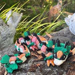 May include: A collection of sushi-themed dragon sculptures. The dragons feature white, orange, and pink sushi rolls with green seaweed accents. The sculptures are arranged on a mossy rock, with a natural background.