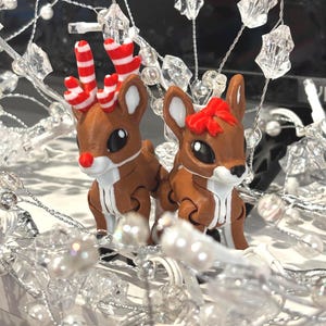 May include: Two brown reindeer figurines with white and red striped antlers and a red nose. One reindeer has a red bow. They are surrounded by silver beaded garland and clear crystal accents, creating a festive holiday scene.