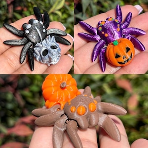 May include: Three 3D printed Halloween spider ornaments. One spider is black with a silver skull, one is purple with a pumpkin, and one is brown with an orange pumpkin.