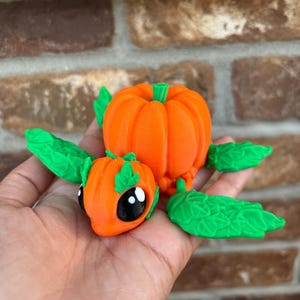 May include: A small, orange pumpkin-shaped figurine with green leaves and black eyes. The figurine is made of a smooth, plastic-like material.