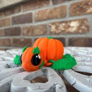 May include: A small, orange pumpkin-shaped figurine with green leaves and black eyes. The figurine is made of plastic and is designed to look like a turtle.