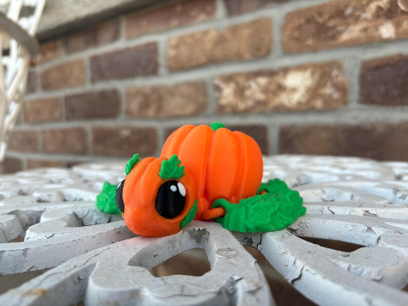 May include: A small, orange pumpkin-shaped figurine with green leaves and black eyes. The figurine is made of plastic and is designed to look like a turtle.