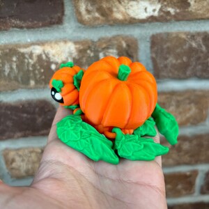 May include: A 3D printed pumpkin figurine with green leaves and a small pumpkin on the side. The pumpkin is orange and the leaves are green.