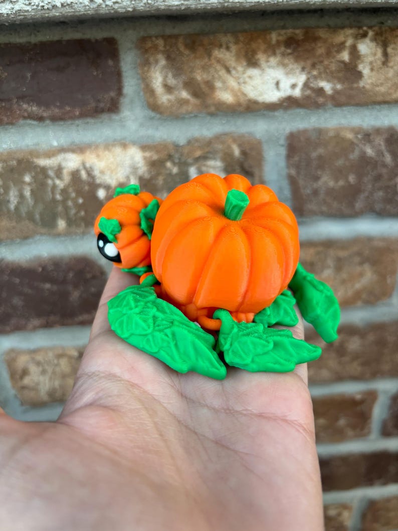 May include: A 3D printed pumpkin figurine with green leaves and a small pumpkin on the side. The pumpkin is orange and the leaves are green.