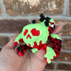 May include: A red and green figurine with a skull-shaped head and spiderweb accents. The figurine has a green, drippy substance over a red skull. Black spiderwebs are on the hands and feet. A black bow is on the head.