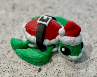 May include: A whimsical green turtle figurine with a red Santa Claus hat and a red shell trimmed with white. The turtle wears a black belt with a white buckle. The figurine is a festive holiday decoration.
