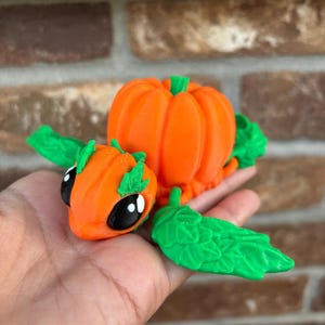 May include: A 3D printed pumpkin-shaped creature with green leaves and black eyes. The creature is orange and has a small pumpkin on its back.