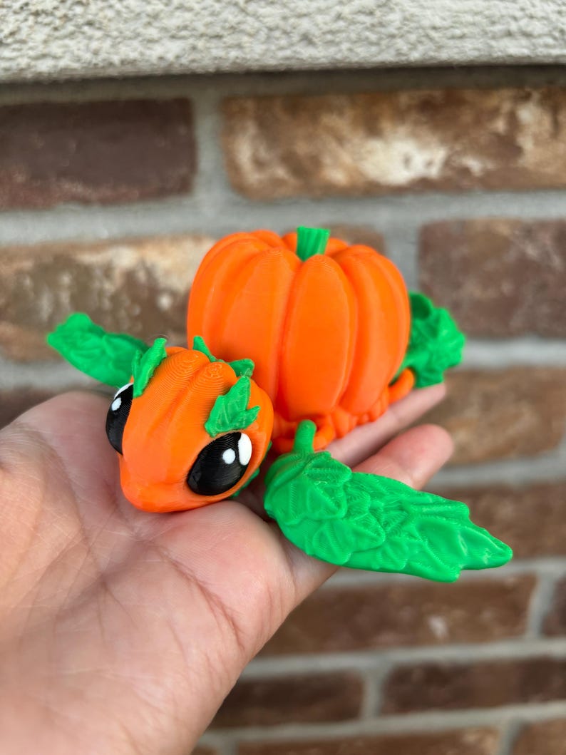 May include: A 3D printed pumpkin-shaped creature with green leaves and black eyes. The creature is orange and has a small pumpkin on its back.