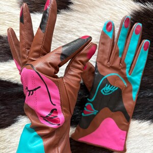 May include: A pair of brown leather gloves featuring abstract face designs in bright pink, turquoise, and black. The gloves have painted fingernail details on the fingertips and bold, colorful graphic patterns across the palms and cuffs.