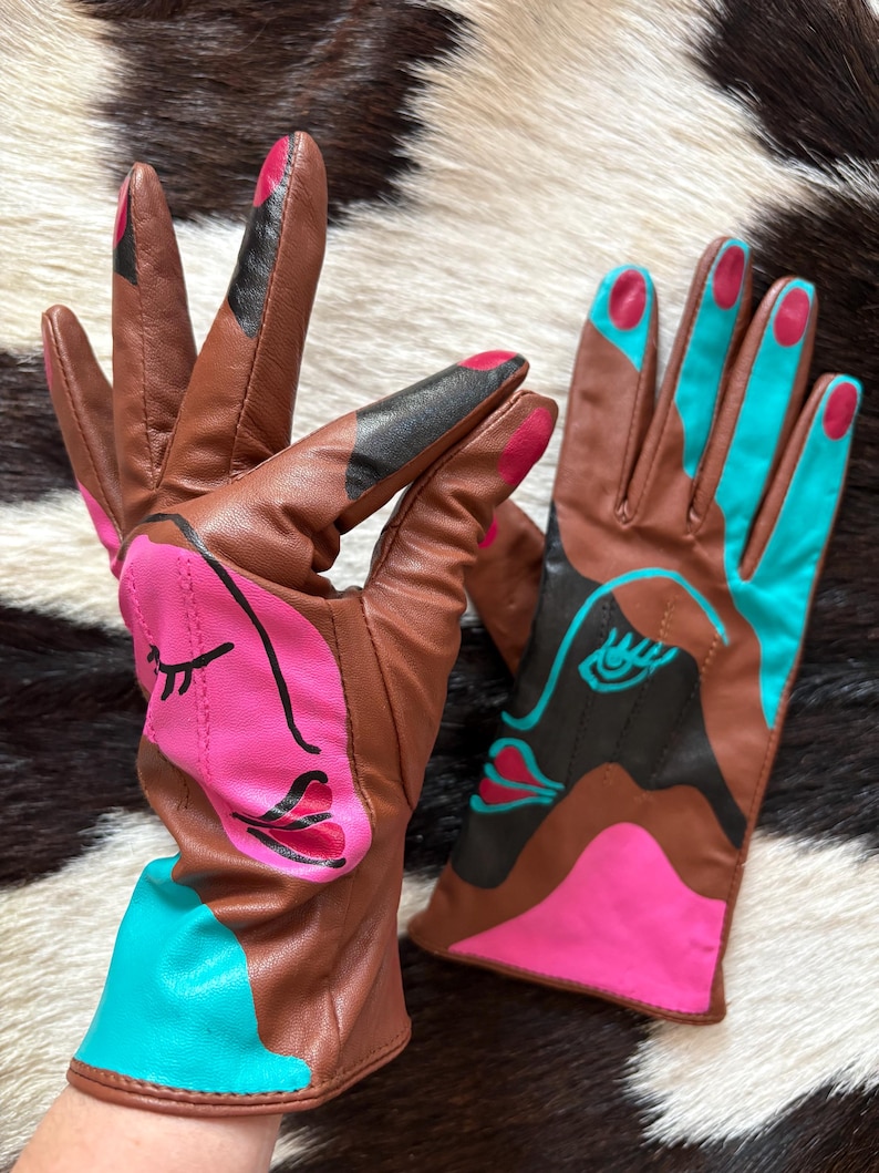 May include: A pair of brown leather gloves featuring abstract face designs in bright pink, turquoise, and black. The gloves have painted fingernail details on the fingertips and bold, colorful graphic patterns across the palms and cuffs.