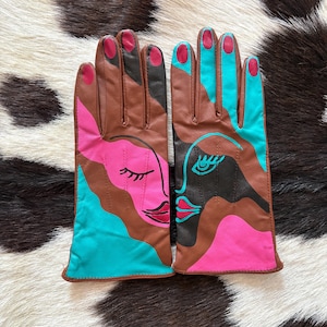 May include: A pair of leather gloves featuring an abstract face design in brown, bright pink, and turquoise. The gloves display stylized facial features including closed eyes, eyelashes, and lips, with pink-painted fingernail details on the fingertips.