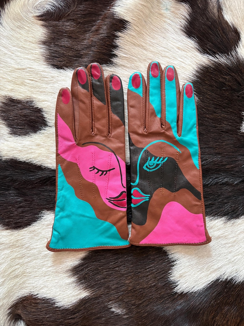 May include: A pair of leather gloves featuring an abstract face design in brown, bright pink, and turquoise. The gloves display stylized facial features including closed eyes, eyelashes, and lips, with pink-painted fingernail details on the fingertips.