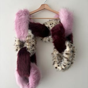 May include: A multi-coloured faux fur scarf featuring an interlocking chain-link design. The scarf is composed of plush segments in solid light pink, deep burgundy, and a cream-coloured faux fur with dark brown leopard-style spots throughout.