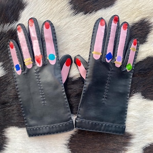 May include: A pair of black leather gloves with painted pink fingernails and red nail polish. Each finger has a gold or jewel-toned ring. The gloves are displayed on a black and white cowhide pattern.