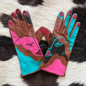 May include: A pair of brown leather gloves featuring abstract face designs in vibrant pink and turquoise. The gloves include painted details such as closed eyes, lips, and colored fingertips, with a smooth finish and visible stitching along the fingers.
