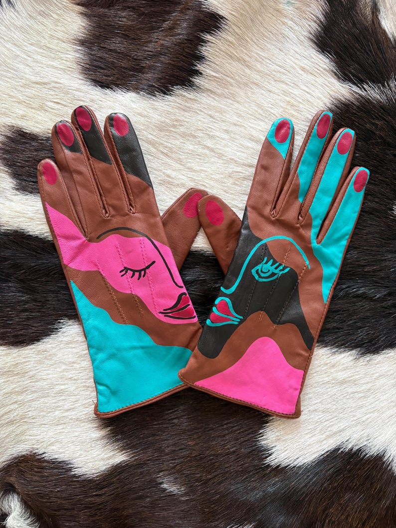 May include: A pair of brown leather gloves featuring abstract face designs in vibrant pink and turquoise. The gloves include painted details such as closed eyes, lips, and colored fingertips, with a smooth finish and visible stitching along the fingers.