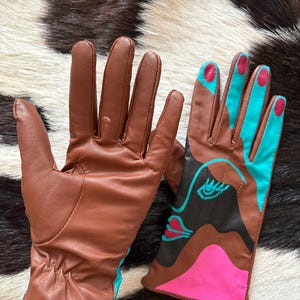 May include: A pair of brown leather gloves. One glove is plain brown, while the other features a graphic design with black, bright pink, and turquoise blue accents, including a stylized eye and lip motif on the back of the hand and colored fingertips.