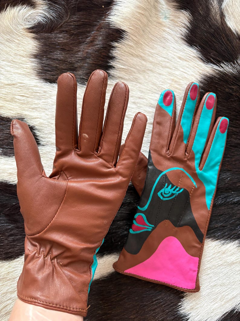 May include: A pair of brown leather gloves. One glove is plain brown, while the other features a graphic design with black, bright pink, and turquoise blue accents, including a stylized eye and lip motif on the back of the hand and colored fingertips.
