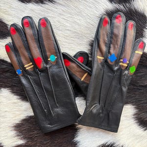 May include: A pair of black leather gloves with painted details. The fingertips are painted red, and the backs of the hands are brown with painted rings in various colours. The gloves are displayed on a black and white cowhide pattern.