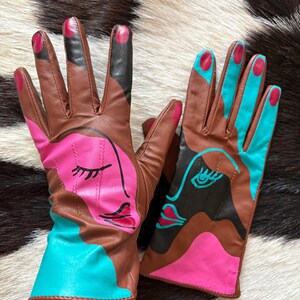 May include: A pair of brown leather gloves featuring abstract face designs in bright pink and turquoise. The left glove displays a pink face with closed eyes, while the right glove shows a turquoise face with an open eye. Both have painted fingertips.