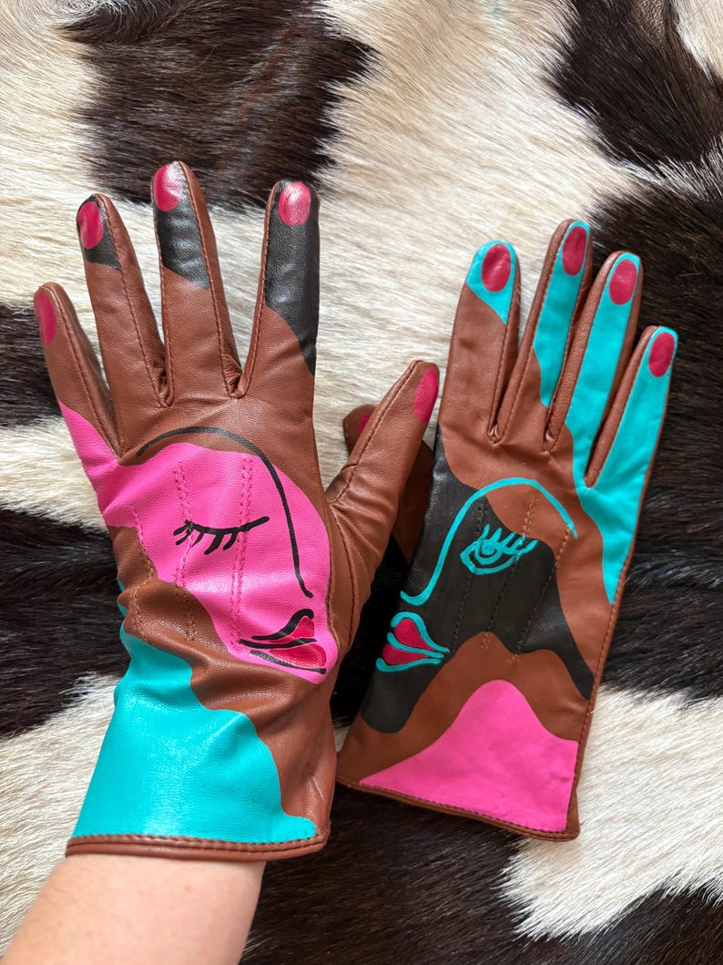 May include: A pair of brown leather gloves featuring abstract face designs in bright pink and turquoise. The left glove displays a pink face with closed eyes, while the right glove shows a turquoise face with an open eye. Both have painted fingertips.