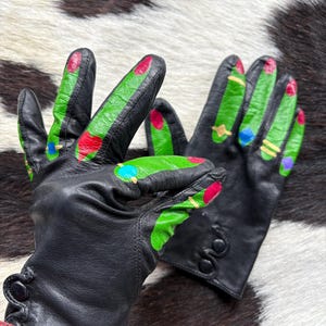 May include: Black leather gloves with painted green fingertips and colorful accents. The gloves have a button detail on the wrist and are set against a cowhide background. The fingertips are painted with red, blue, and yellow accents.