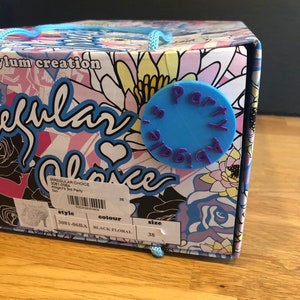 May include: A shoebox with a floral design and a blue circle with the word "Party" in purple lettering. The box has a label with the words "style", "colour", and "size" and the corresponding information: "3081-06BA", "BLACK FLORAL", and "38".
