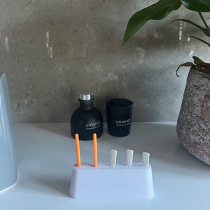 May include: White plastic holder with four orange and white earplugs. The holder is on a white countertop with two black bottles labelled "Unwind" and a grey soap dispenser.