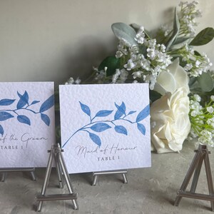 Könnte beinhalten: Zwei weiße Karten mit blauen, blattreichen Zweigen darauf gedruckt. Die Karten stehen auf kleinen Holzstaffeleien. Der Text auf den Karten lautet "Father of the Groom Table 1" und "Maid of Honour Table 1".