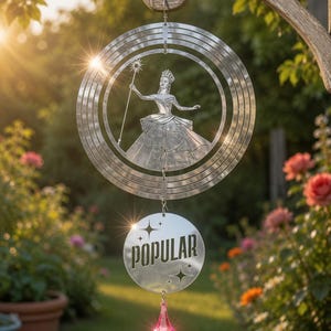 May include: A silver-toned metal wind spinner featuring a circular frame with a cutout of a figure wearing a crown and holding a star-topped wand. Below hangs a circular disc with the word POPULAR and star accents, finished with a pink crystal pendant.