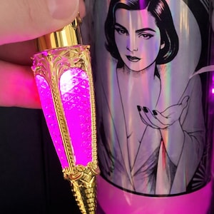 Puede incluir: Un frasco de perfume dorado y ornamentado con un interior rosa brillante se sostiene junto a una botella con el retrato de una mujer. El frasco de perfume tiene un diseño decorativo detallado y una base puntiaguda. La otra botella tiene el texto "IMPRE VIVA".