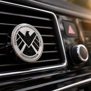 Puede incluir: Un ambientador de coche circular con clip para rejilla de ventilación que presenta un emblema de águila plateada sobre un fondo negro brillante. El texto STRATEGIC HOMELAND INTERVENTION ENFORCEMENT LOGISTICS DIVISION aparece en letras negras.