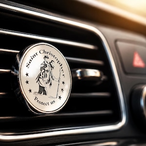 May include: A circular silver-toned car air vent clip featuring a white face with a black illustration of Saint Christopher carrying a child. The text Saint Christopher Protect us is printed in black along the border with small stars.