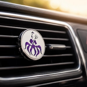 May include: A circular car air freshener vent clip featuring a silver metallic face with a purple glitter graphic of a crowned figure holding a trident. The accessory is attached to the horizontal black slats of a vehicle dashboard air vent.