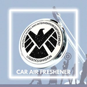 Puede incluir: Un ambientador de coche plateado con un diseño de escudo negro y blanco. El escudo tiene una estrella en el centro y el texto "Strategic Homeland Intervention Enforcement Logistics Division" alrededor.