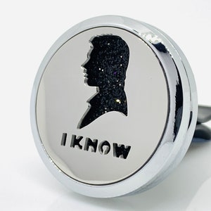 Può includere: Un diffusore per auto in argento con una silhouette nera di una testa di persona e il testo "I KNOW" sotto.