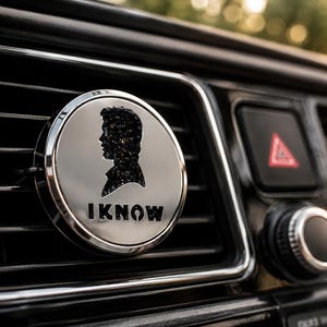 Op de afbeelding: Een ronde zilverkleurige auto-luchtverfrisser voor op het ventilatierooster, voorzien van een zwart silhouet van een persoonlijk profiel met een glittertextuur. De tekst "I KNOW" staat in dikke zwarte hoofdletters onder het silhouet gedrukt.