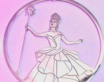 May include: A silver-colored ornament featuring a cutout design of a woman in a gown, holding a staff with a starburst top. The ornament is circular and has a pink ribbon for hanging. The woman is wearing a crown.