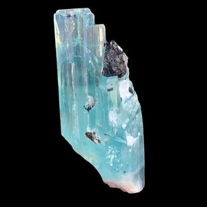 Op de afbeelding: Een doorschijnend, lichtblauw aquamarijnkristal met een donkere, onregelmatige minerale insluitsel. Het kristal heeft een kolomvormige structuur met zichtbare strepen en een enigszins ruwe, gebroken rand. De achtergrond is effen zwart.
