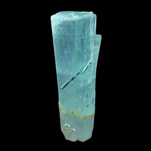 May include: A light blue aquamarine crystal with a clear, glassy appearance. The crystal has a long, slender shape and is slightly translucent. It has a few dark inclusions that run through the crystal.