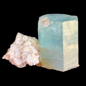 Può includere: Un grande cristallo di acquamarina azzurra di forma rettangolare, accanto a un gruppo di minerali bianchi e beige. L'acquamarina è traslucida, su sfondo nero.