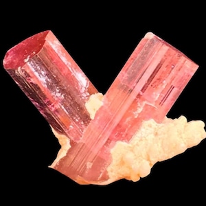 Op de afbeelding: Een close-up van een roze toermalijnkristalformatie. Twee langwerpige, doorschijnende kristallen kruisen elkaar en tonen een reeks roze tinten. De kristallen rusten op een basis van gebroken wit, getextureerd materiaal tegen een zwarte achtergrond.