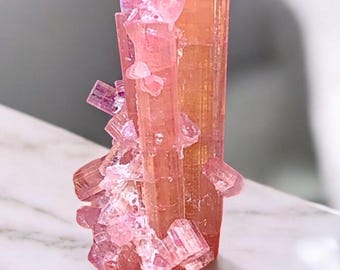 May include: A pink tourmaline crystal cluster. The crystal has a columnar structure with a translucent appearance and a range of pink hues. Smaller crystal formations are attached to the main body, creating a textured surface. The crystal sits on a white marble surface.
