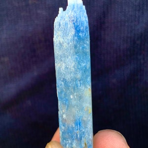 Op de afbeelding: Een lichtblauw kristal met een ruw, getextureerd oppervlak. Het kristal is lang en dun, met een puntige punt.