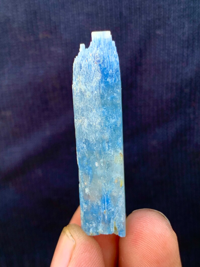 Op de afbeelding: Een lichtblauw kristal met een ruw, getextureerd oppervlak. Het kristal is lang en dun, met een puntige punt.