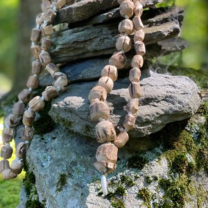 May include: A long strand of wooden beads, strung together with white cord, is draped over a stack of grey rocks. The beads are a light brown colour and have a natural, rustic look.