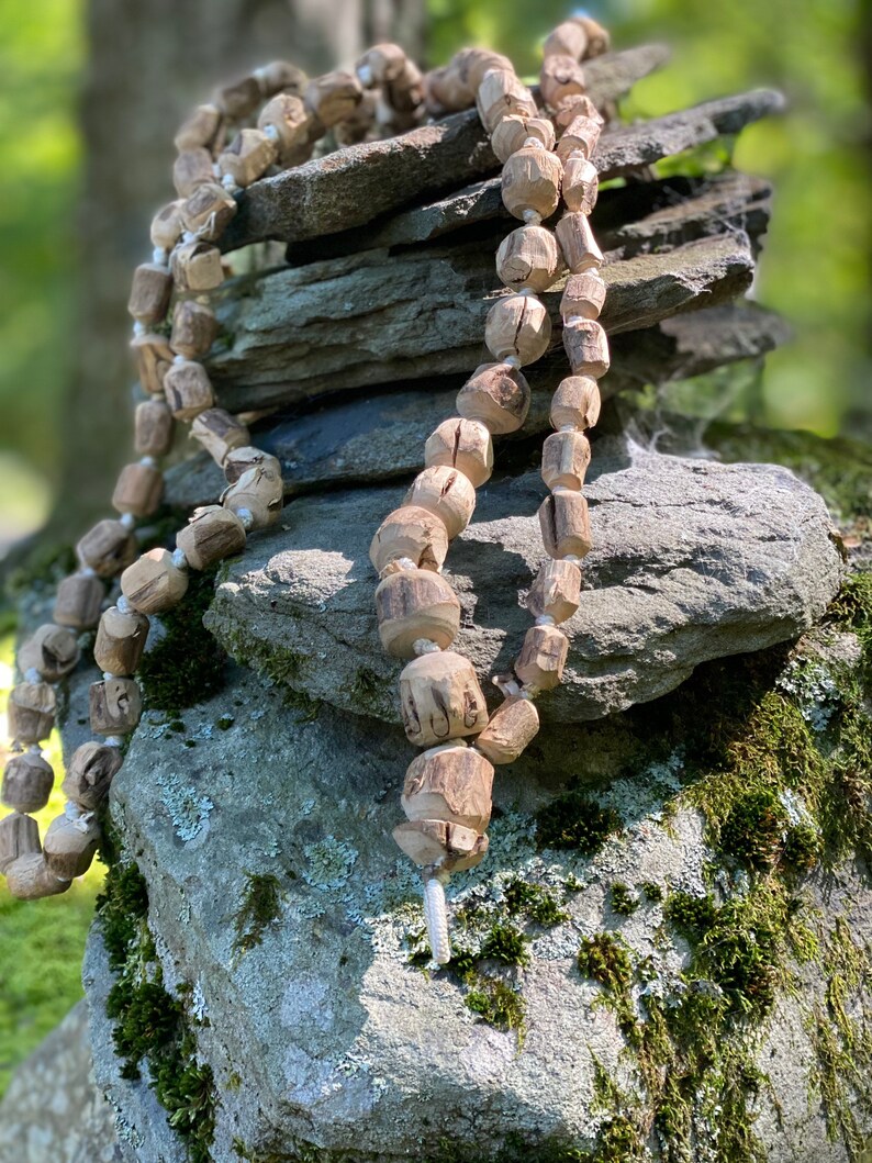May include: A long strand of wooden beads, strung together with white cord, is draped over a stack of grey rocks. The beads are a light brown colour and have a natural, rustic look.
