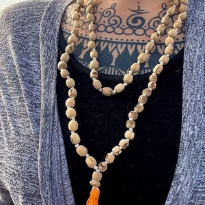 May include: A long, brown wooden beaded necklace with a small orange tassel. The necklace is worn by a person with a black tattoo on their chest.