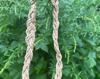 May include: A beige braided necklace made of small, round beads. The necklace is hanging from a green leafy branch.