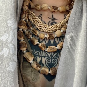 May include: A layered necklace made of multiple strands of wooden beads. The beads are a light brown colour and have a natural, rustic look. The necklace is worn over a brown shirt and a grey scarf.